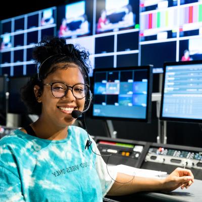 A young woman with glasses and a headset, smiling while sitting in front of a control panel in a media production room. Multiple screens showing various video feeds are visible in the background.